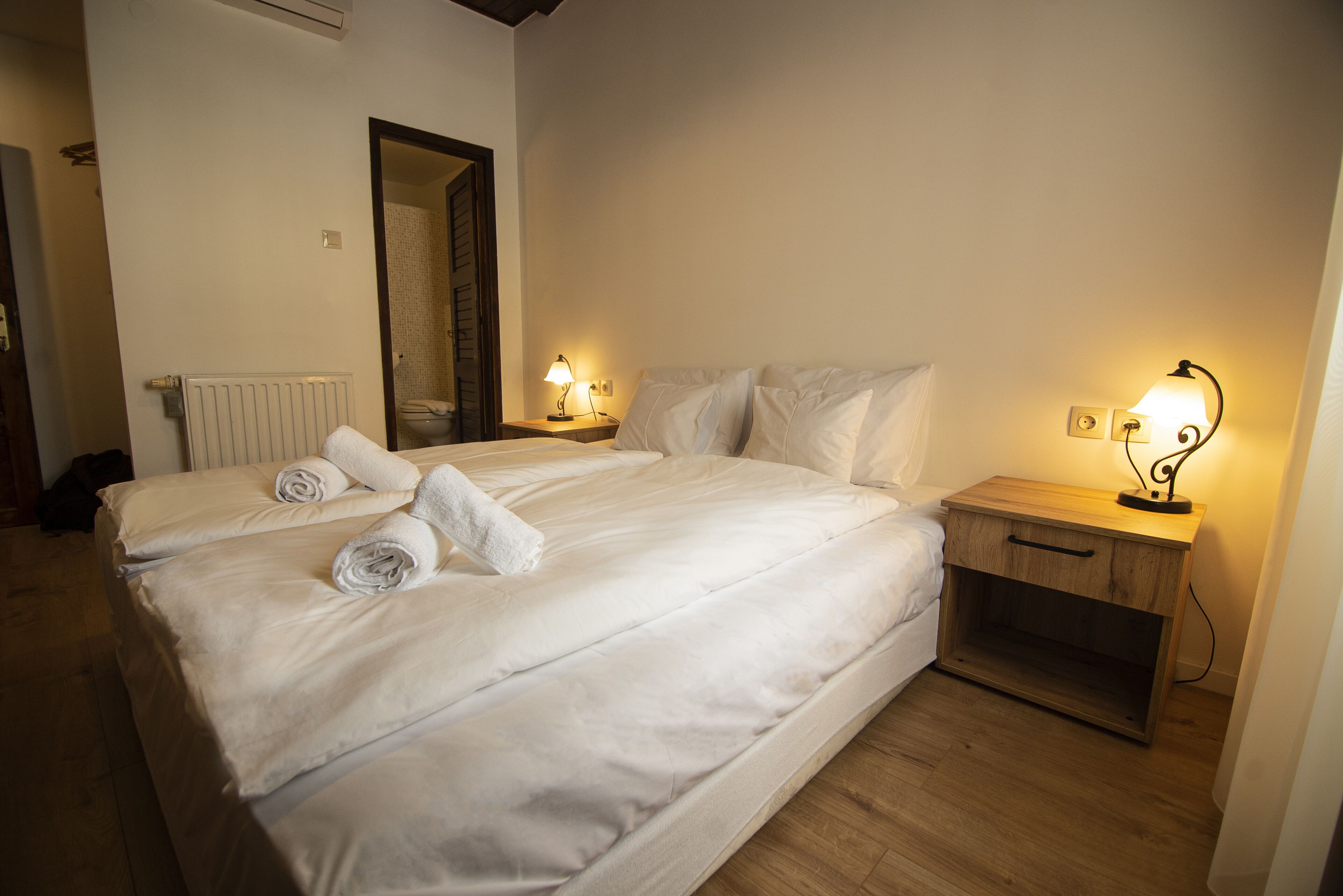 A double bed with white linens and rolled towels, two bedside tables with lamps, a wooden nightstand, and a radiator.