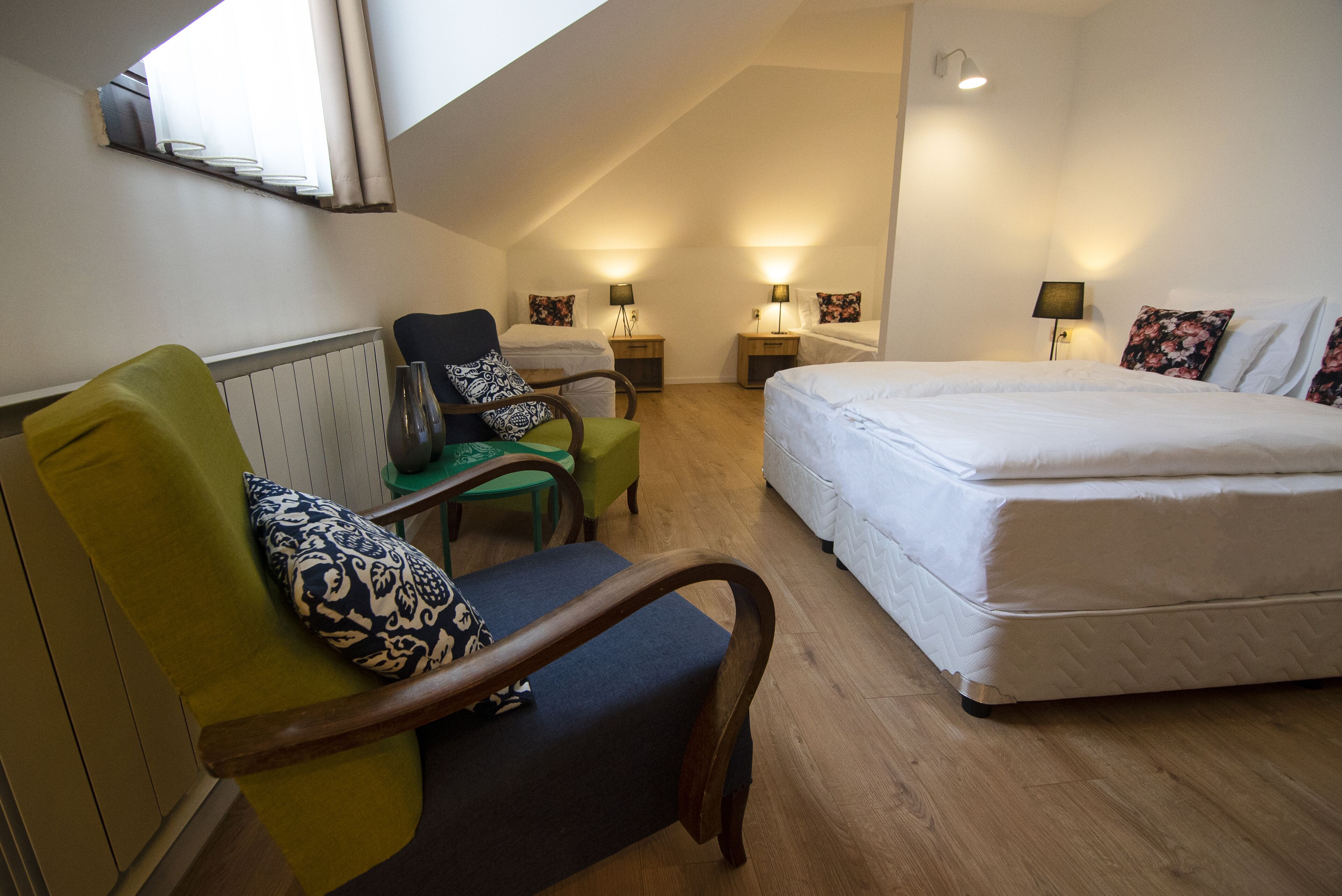 A hotel room with a large bed, a green armchair with a patterned pillow, a small table, and two bedside tables with lamps.
