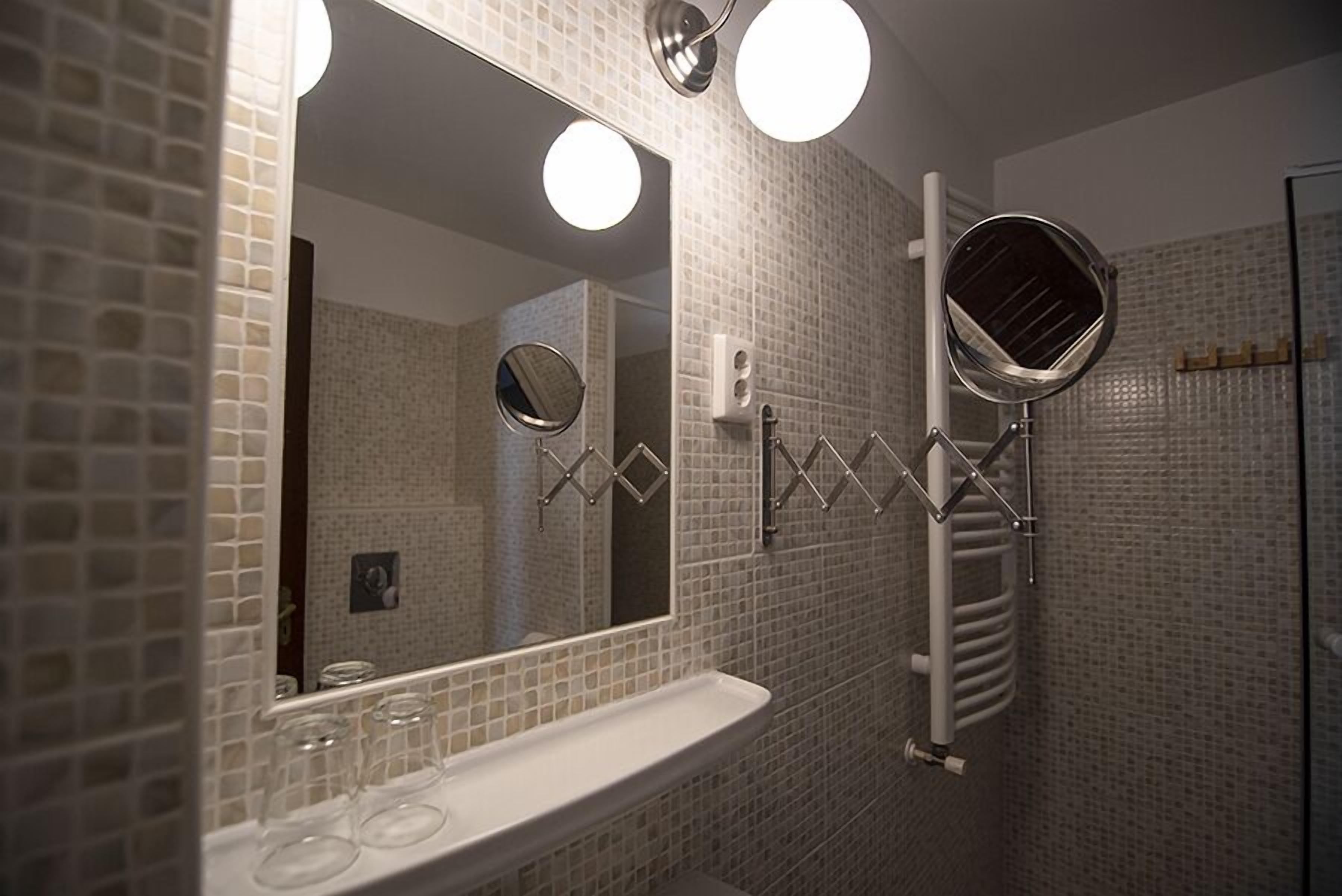 A bathroom with a tiled wall, a sink with a mirror, and a hairdryer mounted on the wall.