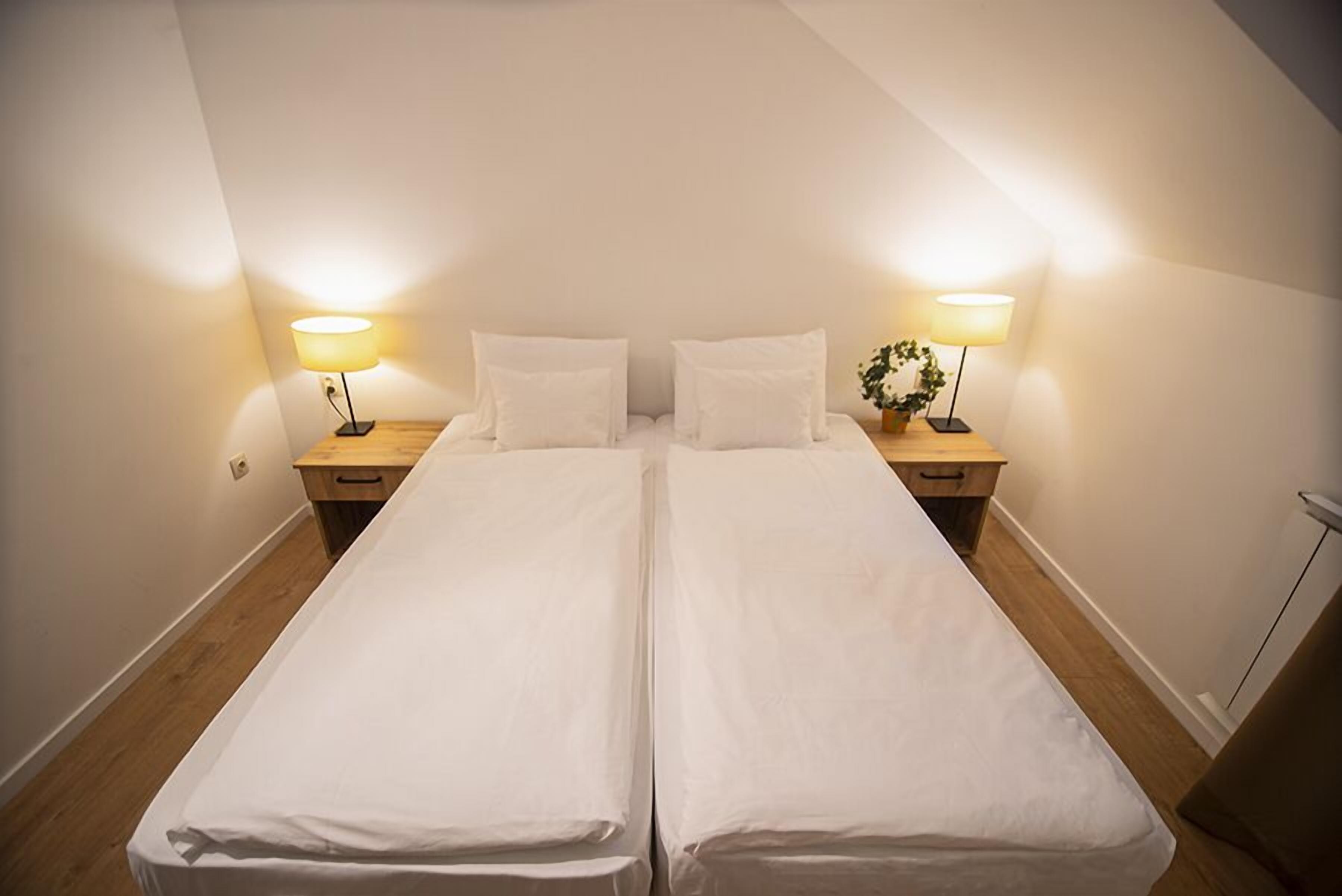 Habitación con dos camas individuales, dos mesitas de noche, cada una con lámpara, paredes blancas y suelo de madera, todo en un ambiente sencillo y limpio.