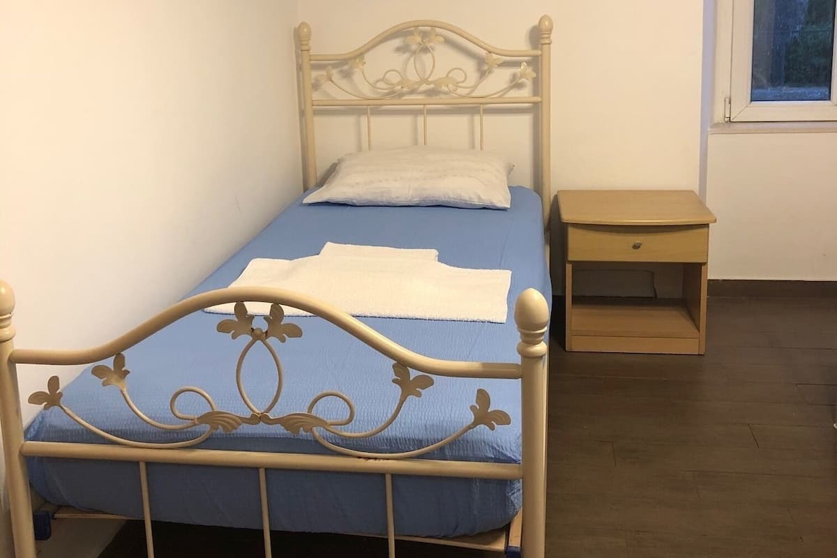 Una cama individual con estructura metálica y colchón azul, una mesita de noche y una ventana con vistas al exterior.