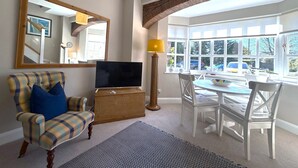 A living room with a television, a plaid armchair, a dining table with chairs, and a large window with a view of the outside.