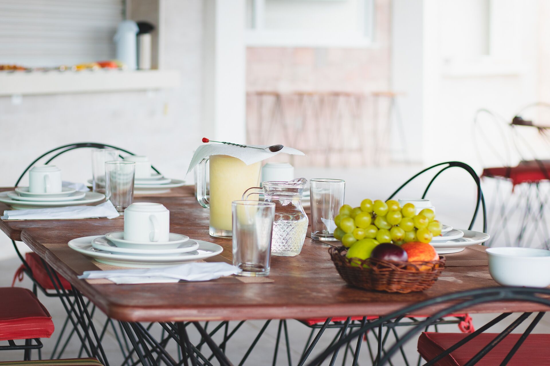 Une table à manger dressée avec des assiettes, des verres, un pichet et un panier de fruits.