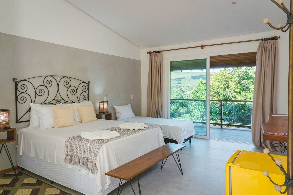 Ein Schlafzimmer mit zwei Betten, einer Bank, einem Nachttisch mit Lampe und Blick auf Grün hinter einer Schiebetür.