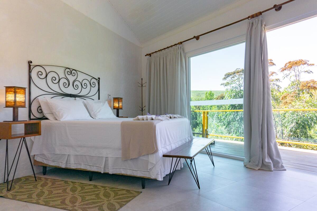 Ein Schlafzimmer mit Bett, Nachttischlampen, einer Bank und einem Balkon mit Vorhängen.