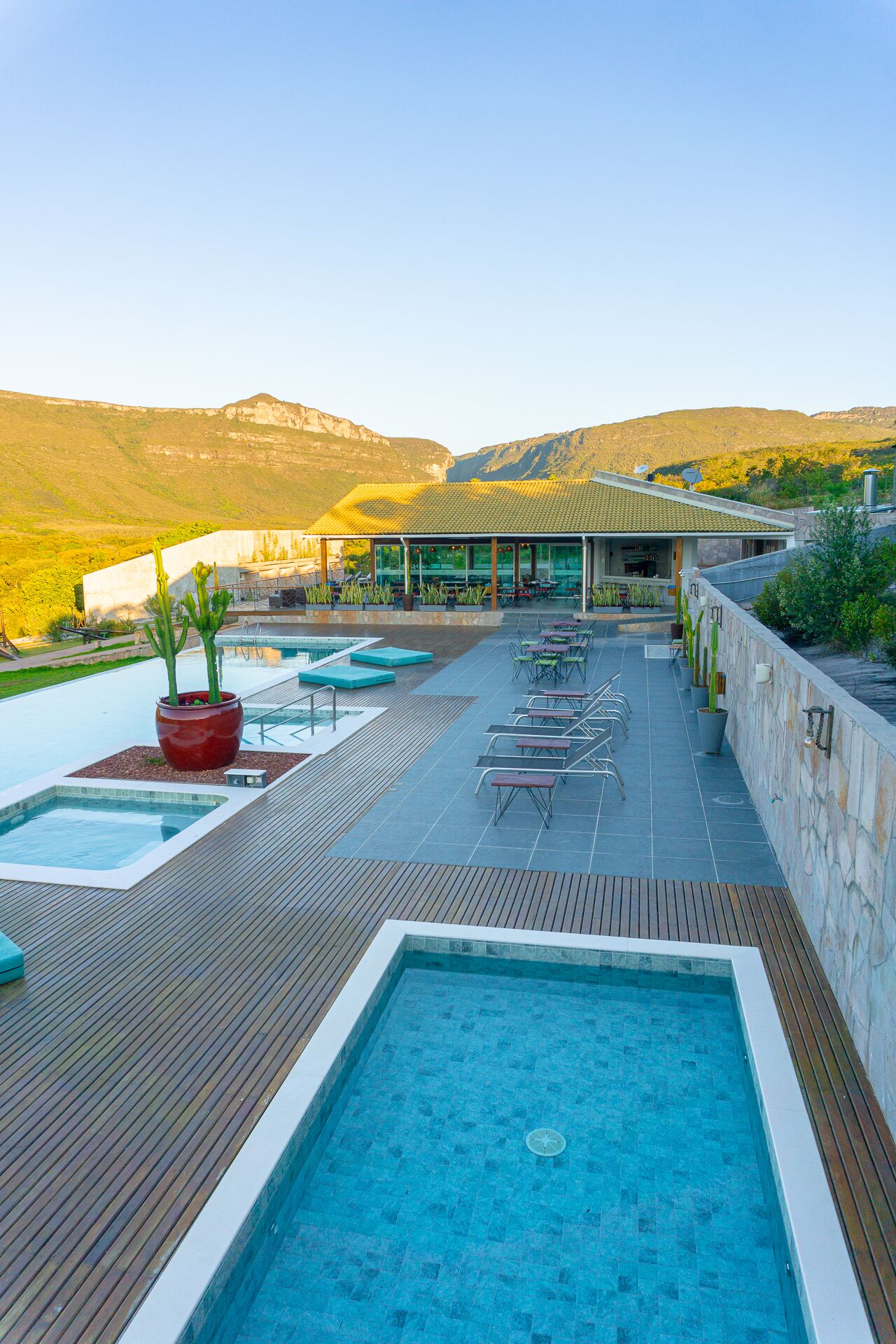A pool area with multiple pools and lounge chairs, surrounded by a wooden deck and stone walls, with a mountain backdrop.