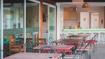 An indoor dining area with tables and chairs, a kitchen in the background, and a glass wall with decorative elements.