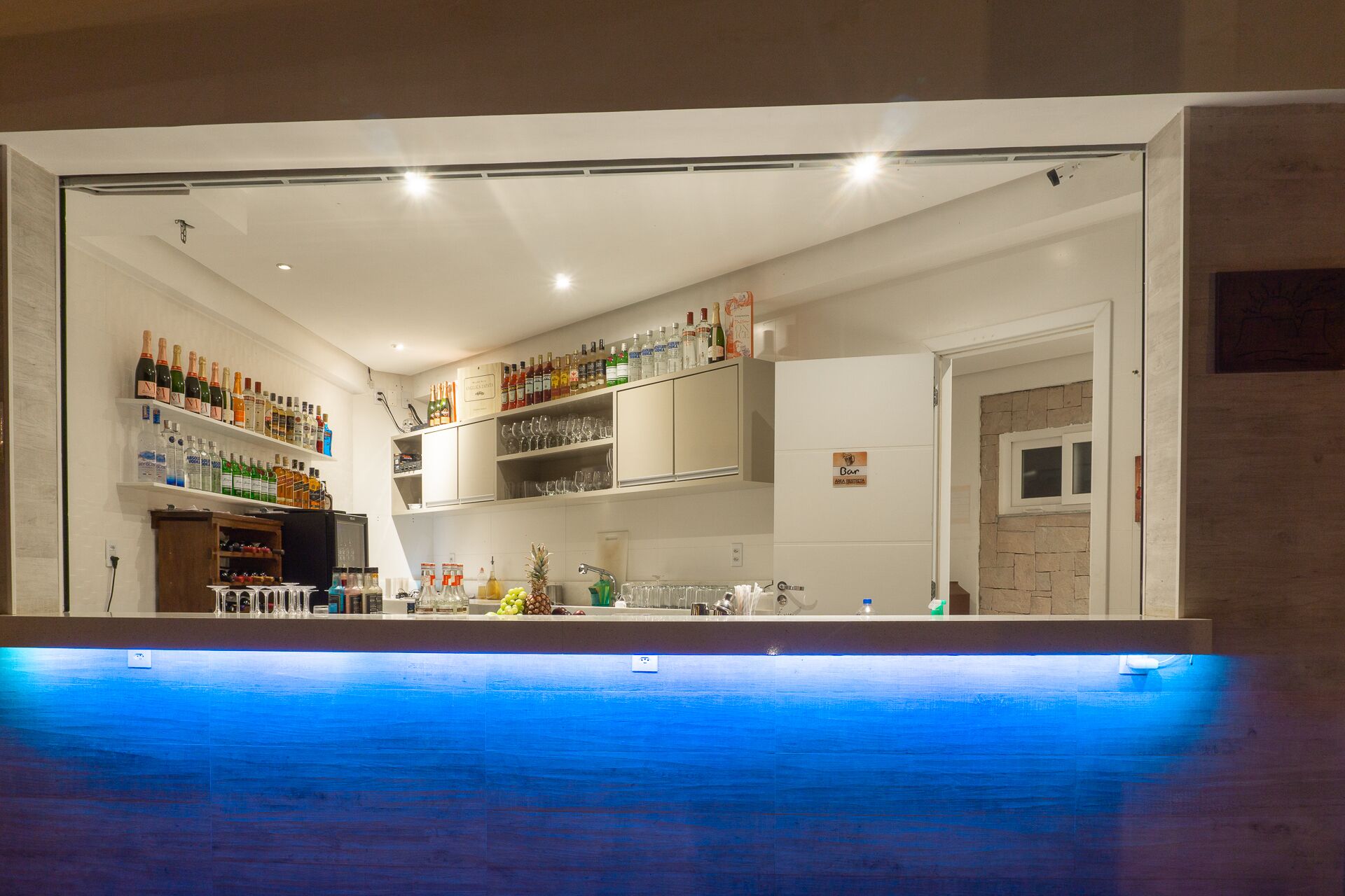 A bar area with shelves of bottles and glasses, a counter with a blue-tiled floor, and a stone wall in the background.