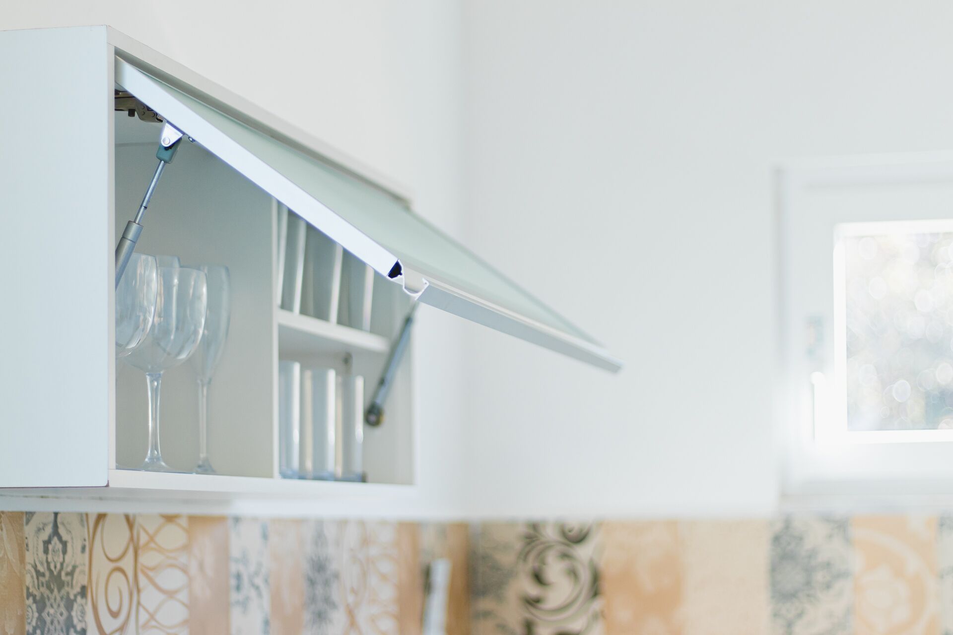 A kitchen with a white cabinet, a hanging rack with wine glasses, and patterned backsplash tiles.