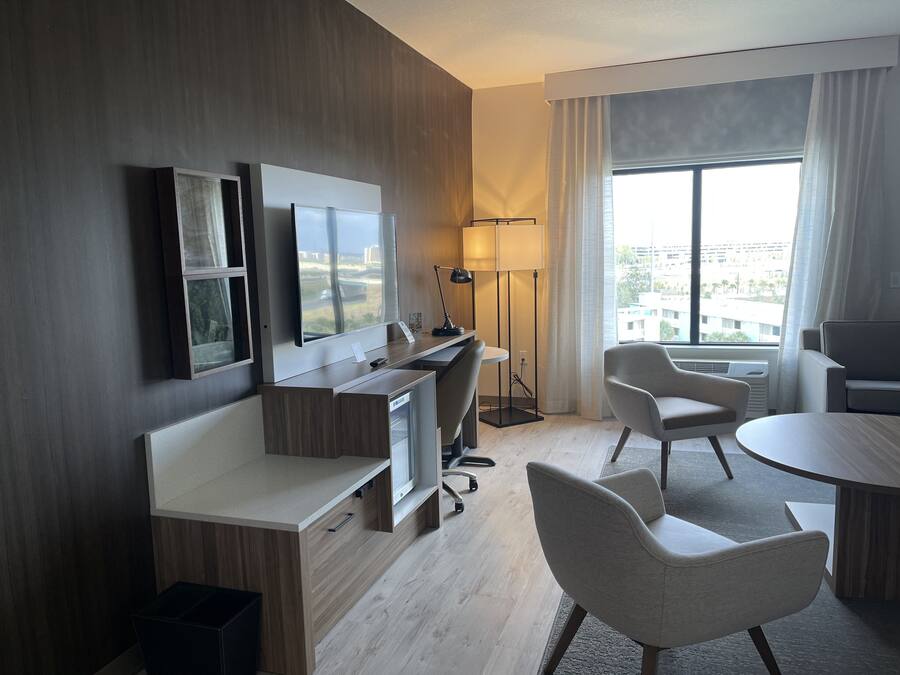 Ein modernes Hotelzimmer mit Schreibtisch aus Holz, zwei Stühlen, einem Flachbildfernseher und einem großen Fenster mit Blick auf die Stadt.