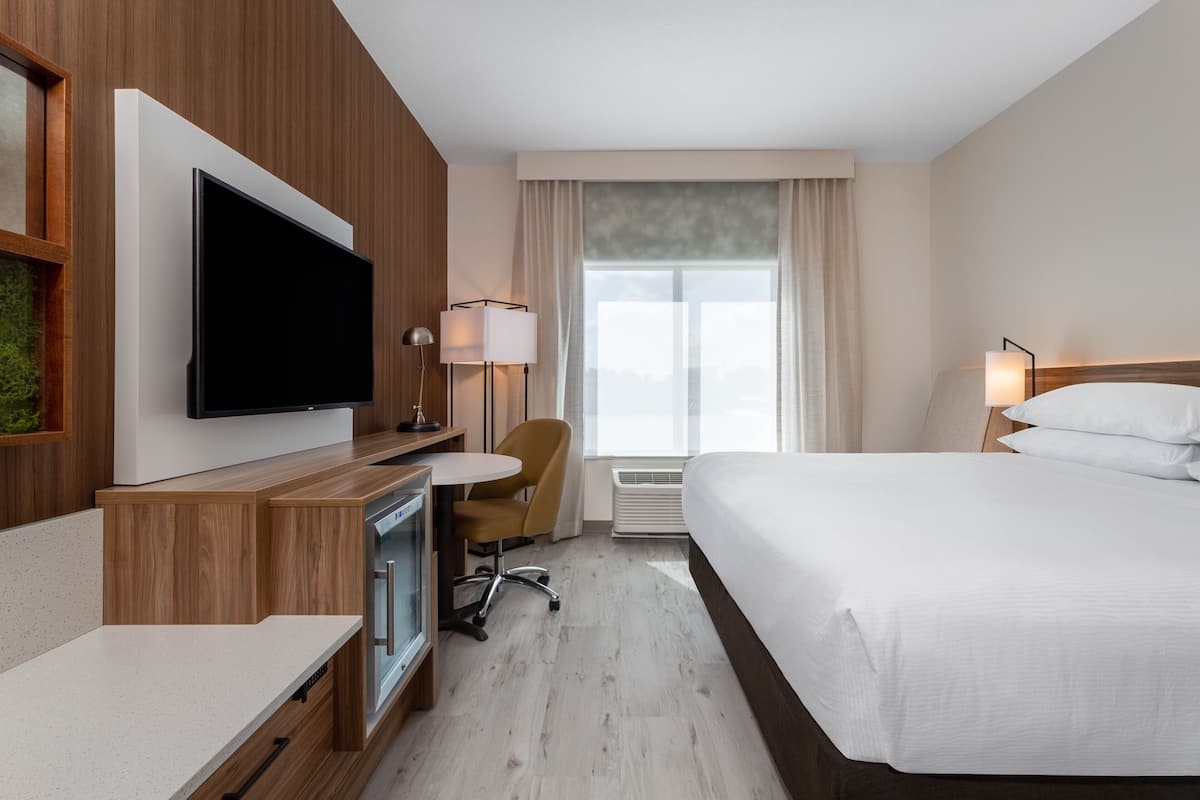 Habitación de hotel con cama, escritorio con televisor, silla, mesita y ventana con cortinas.