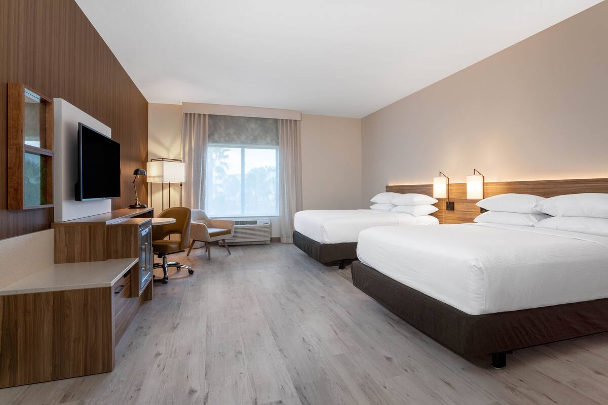 Habitación de hotel con escritorio, dos camas, televisión y ventana con cortinas.