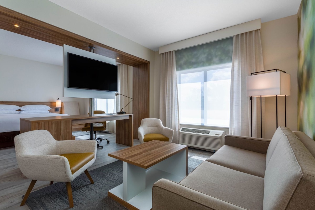 Habitación de hotel con cama, escritorio, televisor, sofá, sillón, mesa de centro y un gran ventanal con cortinas.