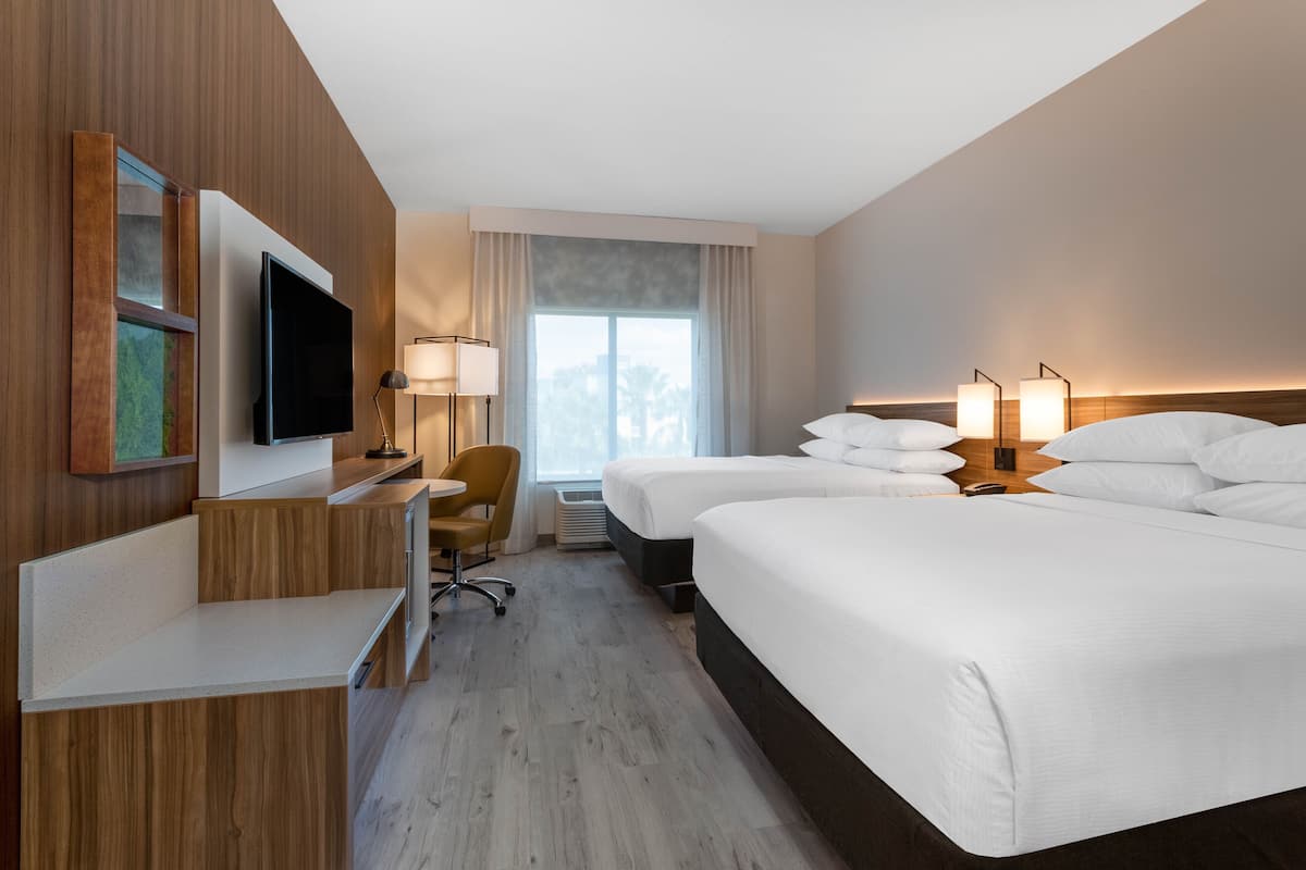 Habitación de hotel con televisión de pantalla plana, escritorio, dos camas, una silla y un ventanal con cortinas.