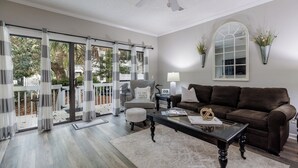 A living room with a brown leather sofa, a coffee table, and a chair. There is a window with curtains, a ceiling fan, and a wall-mounted lamp. - 46 Ocean Breeze: Luxury Villa, Newly Updated, Pet Friendly, Free Bikes (Hilton Head Island)