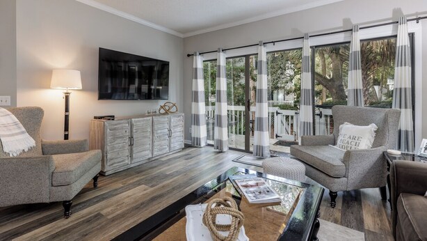 A modern living room with a large TV, comfortable seating, a glass coffee table, and a view of the outdoors through large windows. - 46 Ocean Breeze: Luxury Villa, Newly Updated, Pet Friendly, Free Bikes (Hilton Head Island)