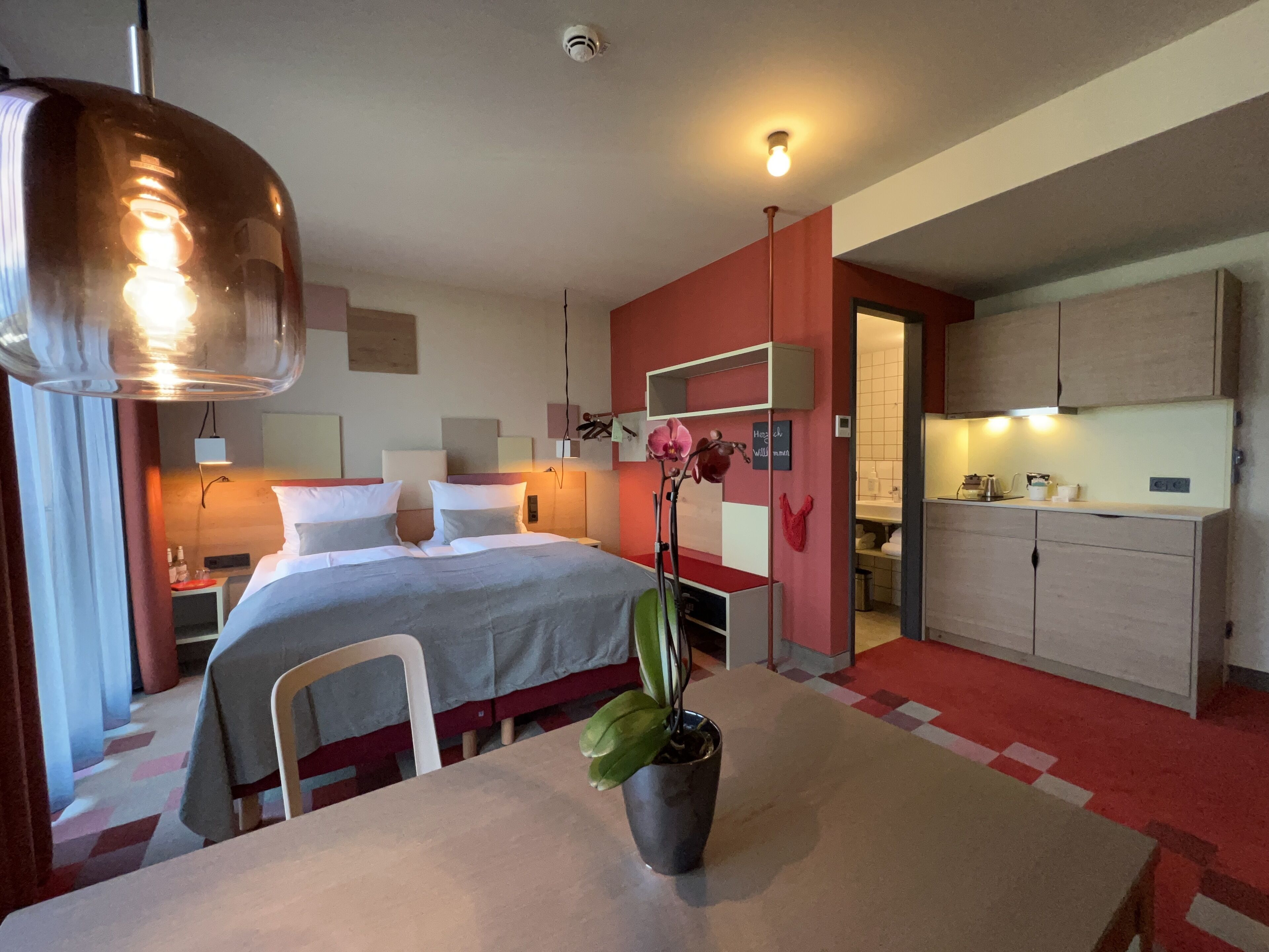 Una habitación de hotel moderna con cama, mesitas de noche, escritorio, mesa de comedor y zona de cocina.