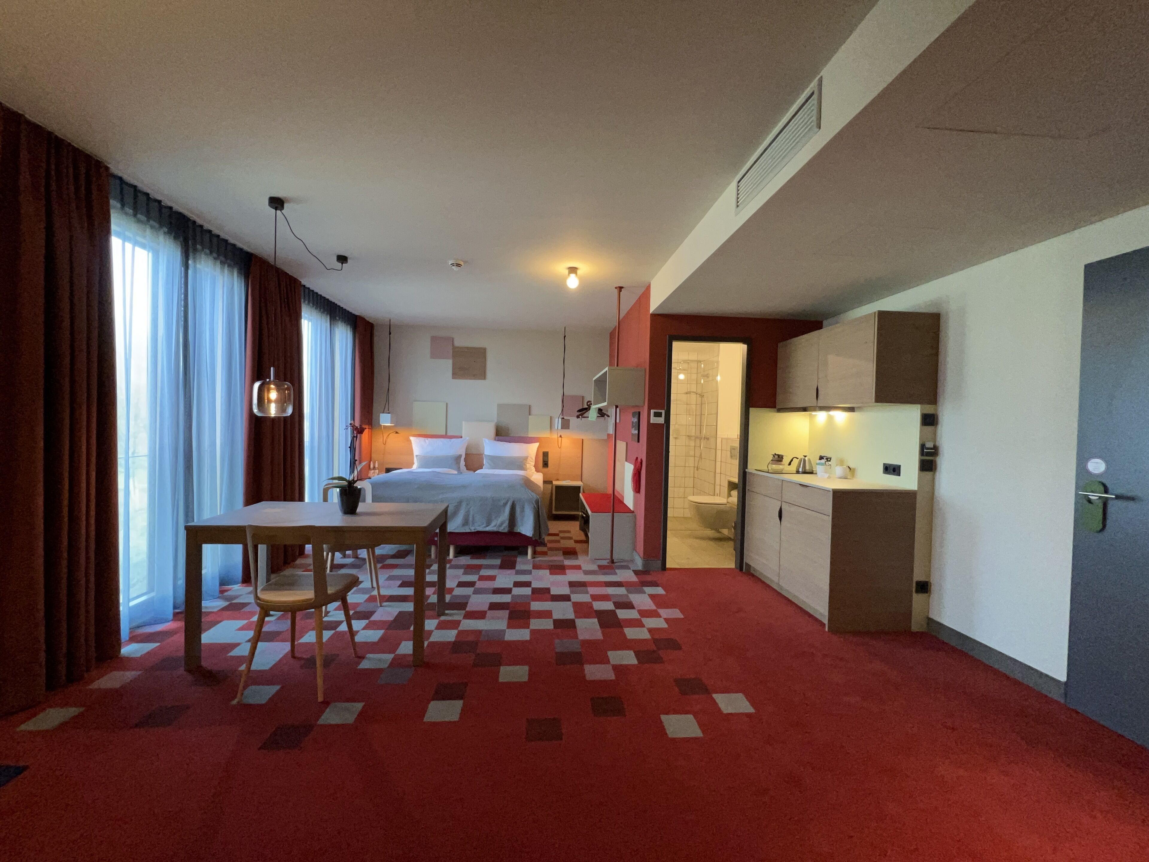 A hotel room with a bed, a desk, a chair, a kitchenette, and a bathroom.