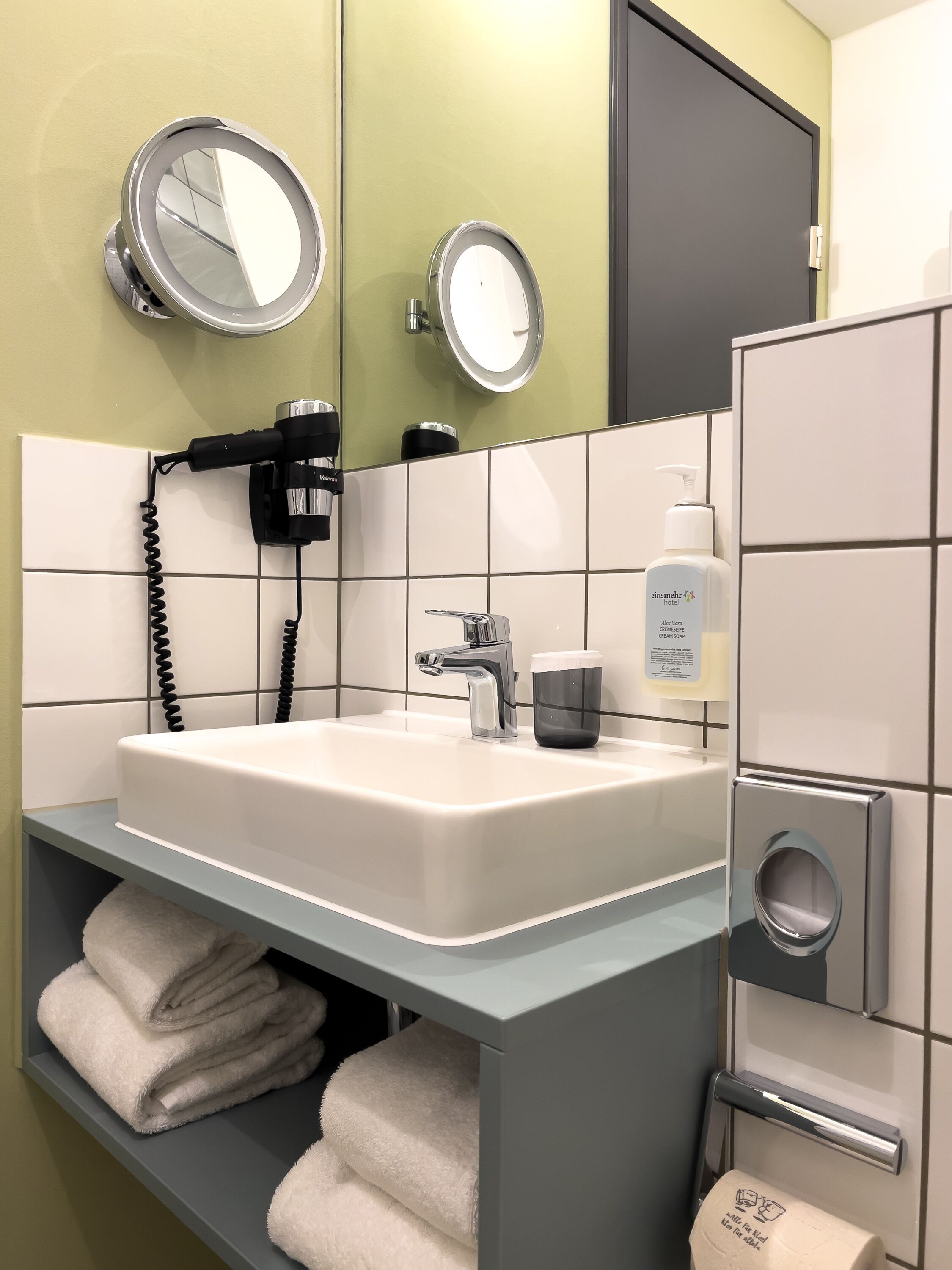 A modern bathroom with a white sink, two round mirrors, a hairdryer, a bottle of aloe vera cream, and neatly folded towels on a shelf.
