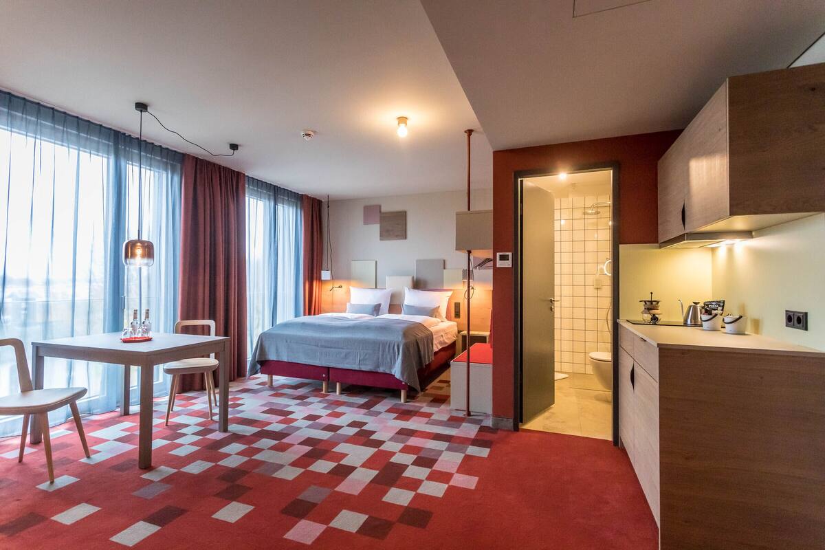 Une chambre d’hôtel comprenant un lit, un bureau, une chaise, un tapis à carreaux rouges et blancs, une salle de bain avec des toilettes et un lavabo, et une kitchenette équipée d’une cuisinière et de placards.