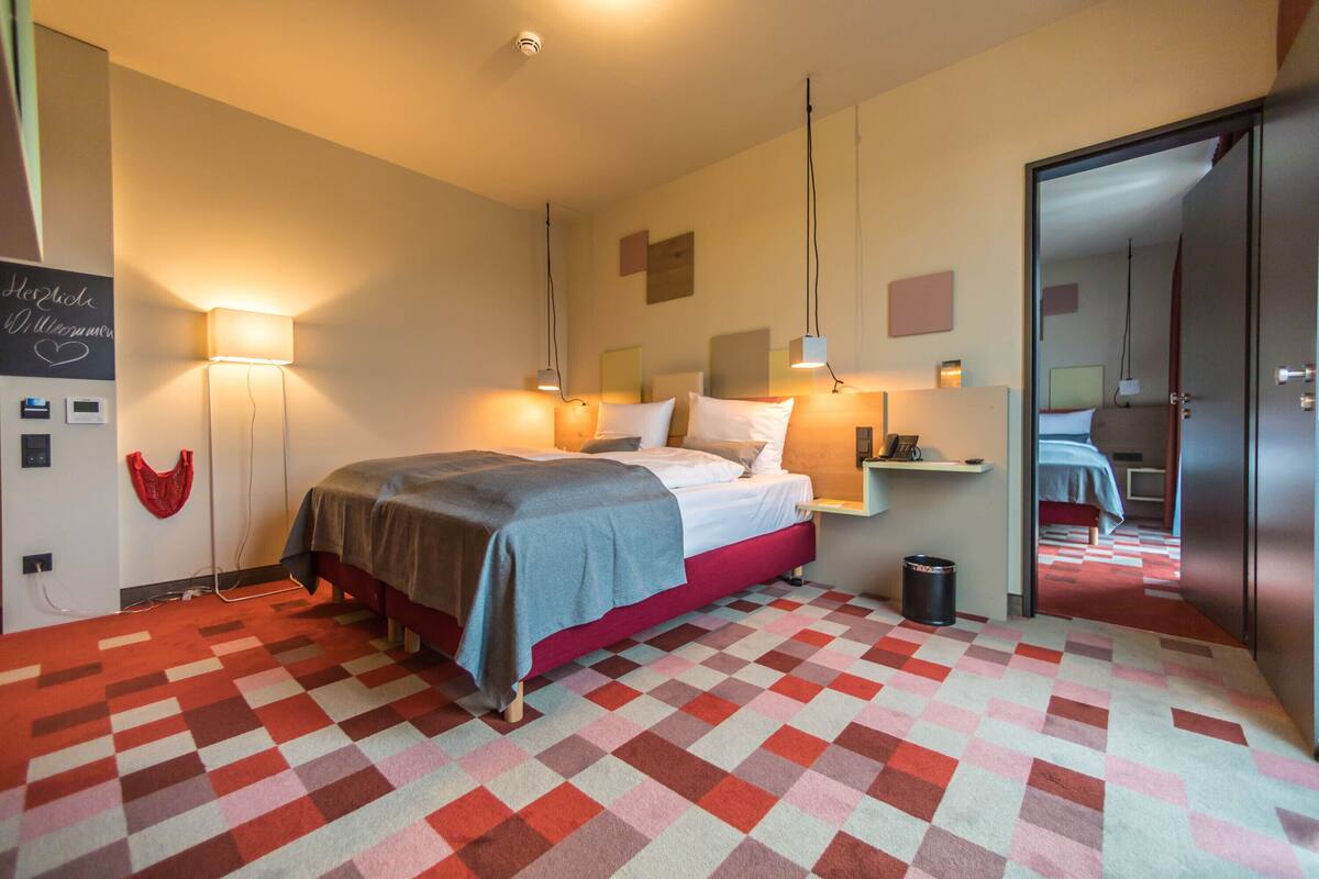 Une chambre d’hôtel avec un tapis à carreaux, un lit recouvert d’une couverture grise, des lampes de chevet et une petite poubelle.