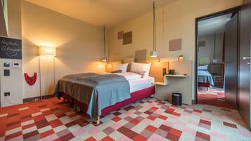 Una habitación de hotel con alfombra a cuadros, una cama con manta gris, lámparas de noche y un pequeño cubo de basura.