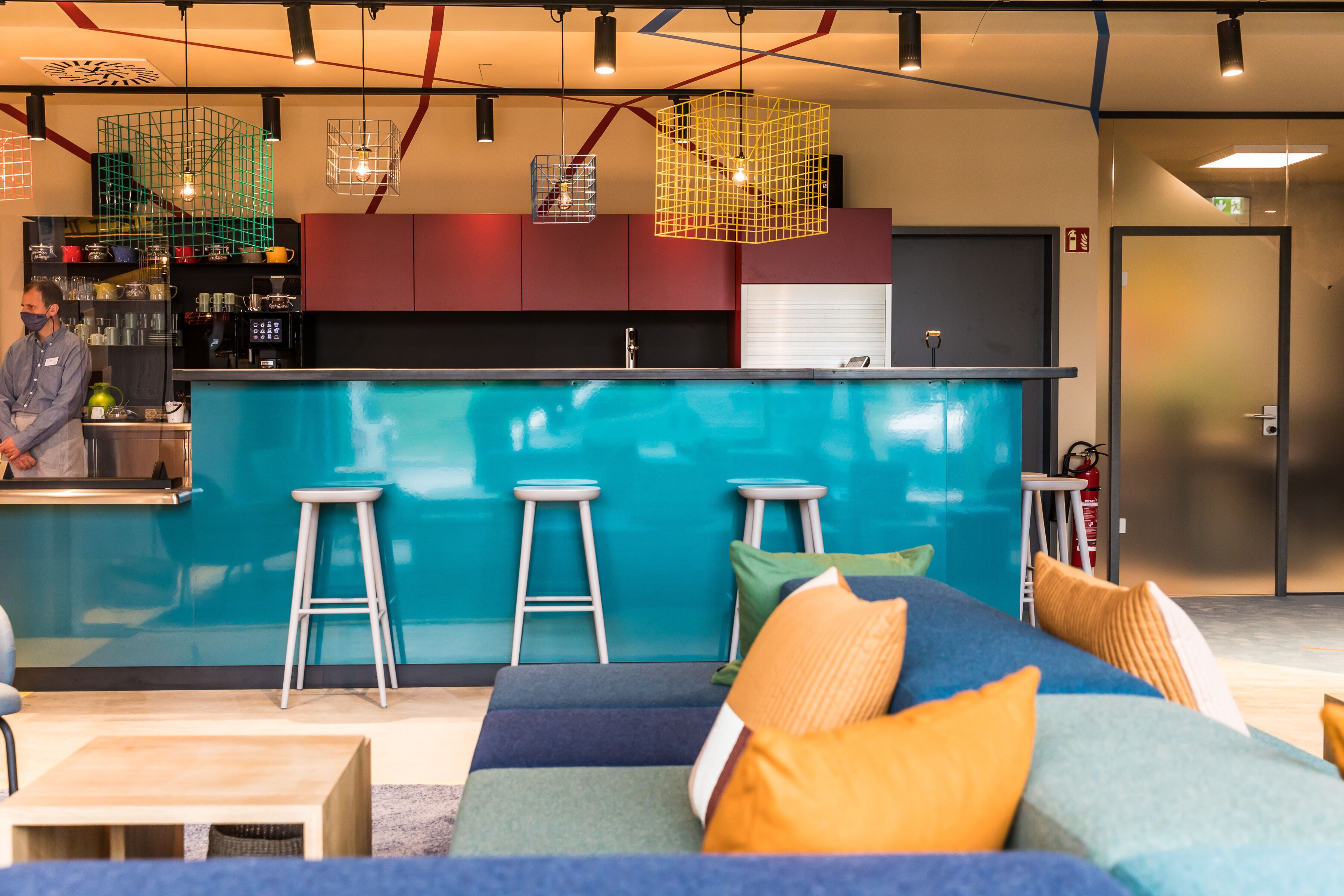 Un'area bar moderna con un bancone blu, tre sgabelli e un angolo salotto con cuscini colorati.