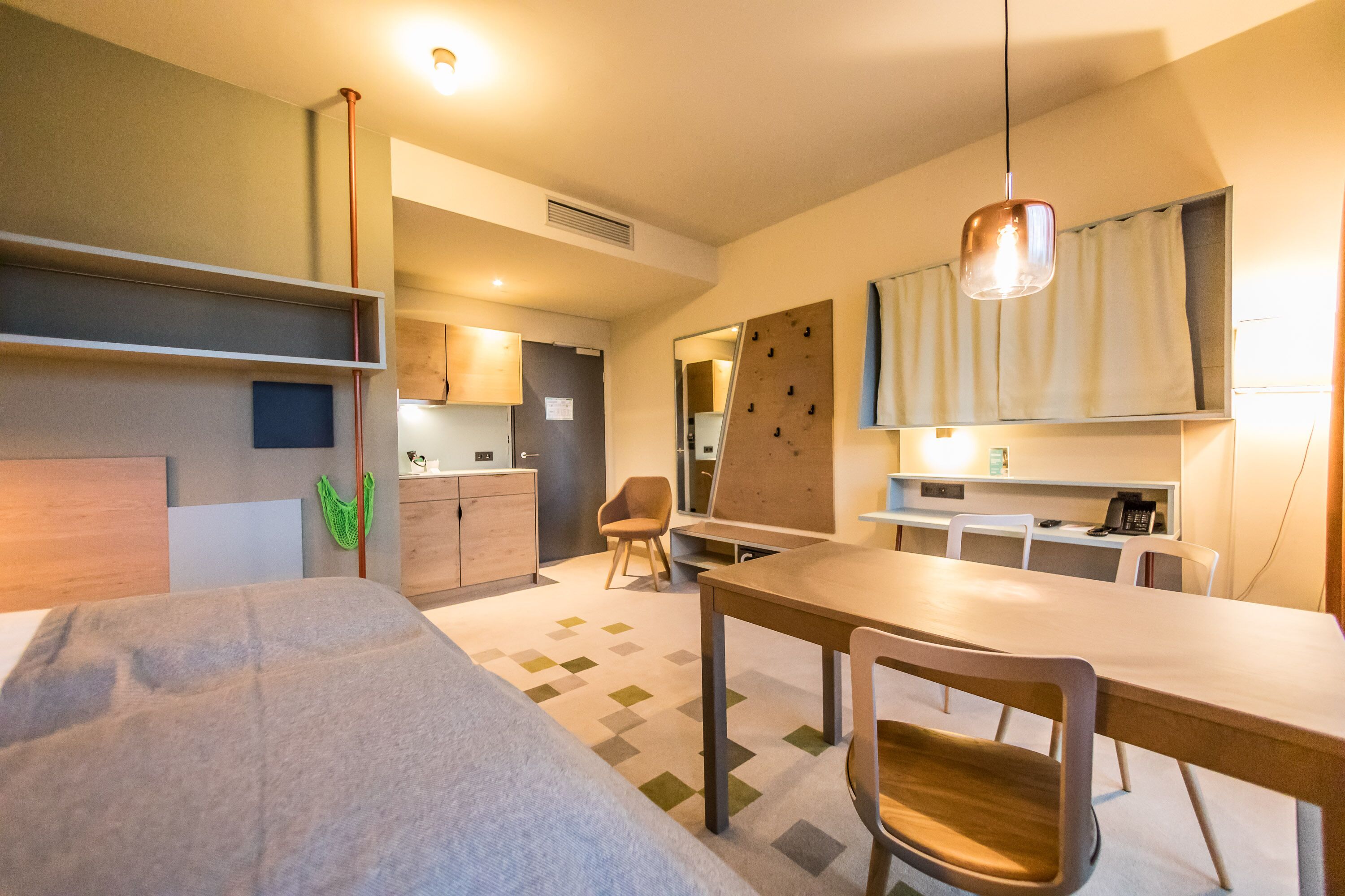 Una habitación de hotel moderna con cama, una mesa de comedor, una silla, una pequeña cocina y un espejo.