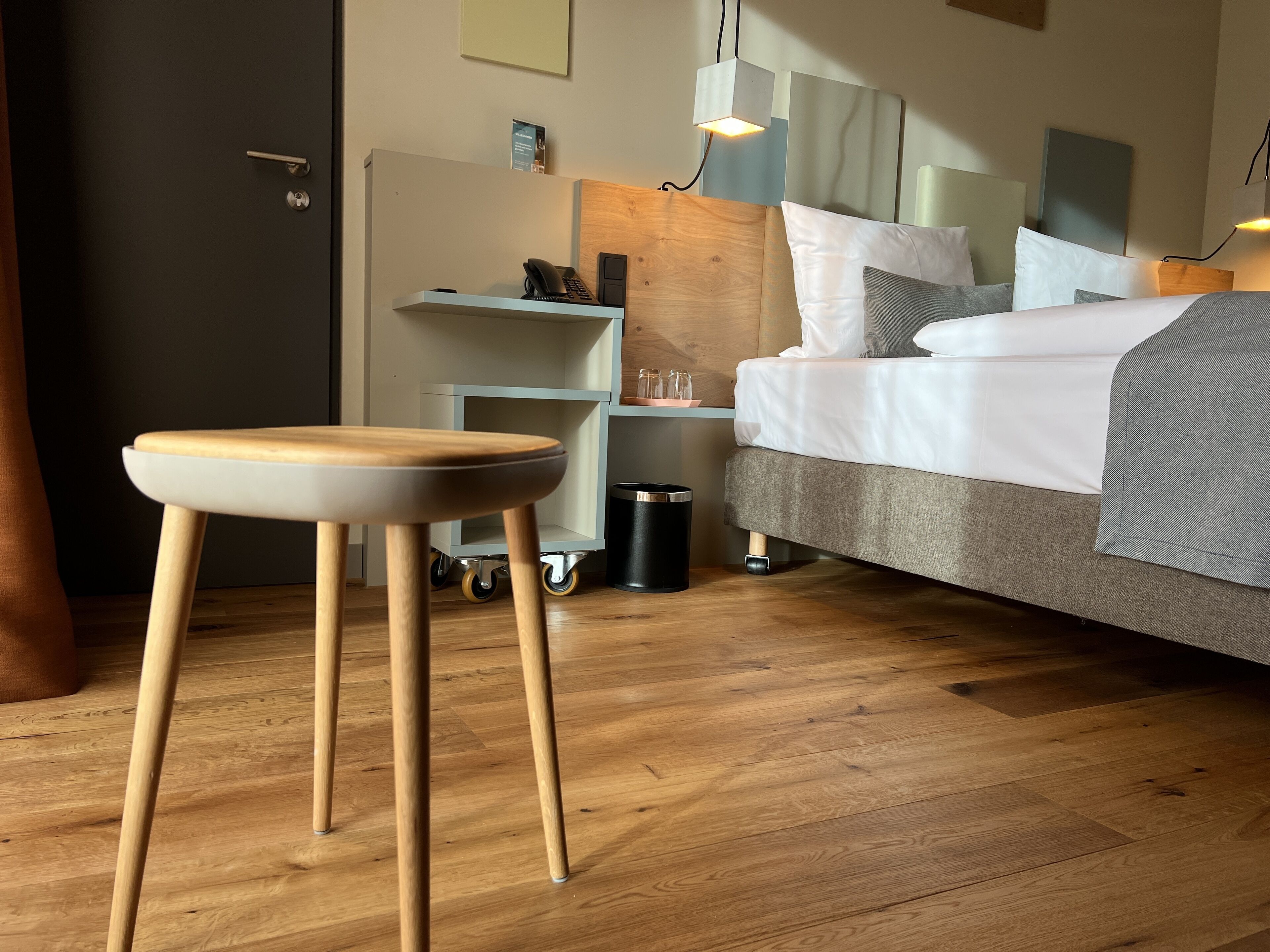 A modern hotel room with a wooden floor, a bed with white and gray bedding, a small desk with a phone, and a trash bin.