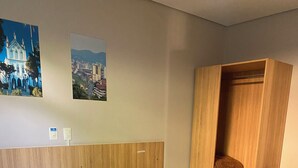 A hotel room with a bed, a wooden wardrobe, a chair, and two wall-mounted light switches. - Hotel Mime (Blumenau)