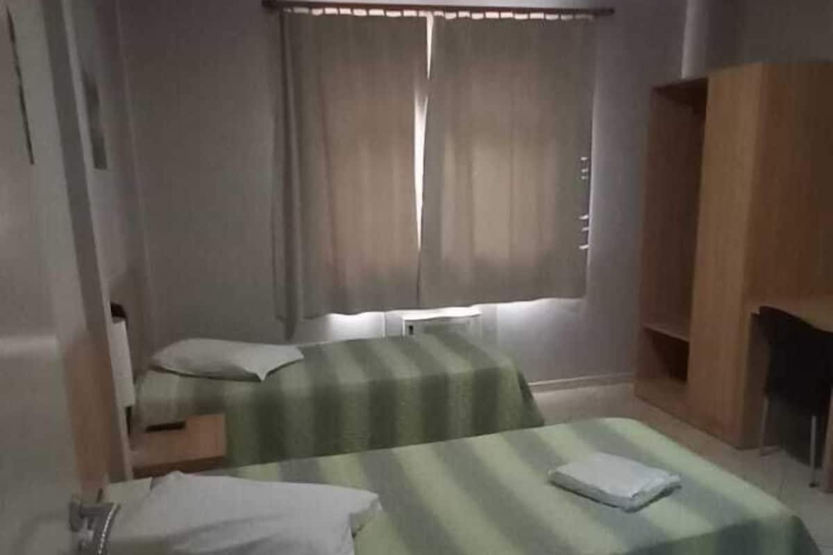 A hotel room with two beds, a small table, a chair, a window with curtains, and a tiled floor.