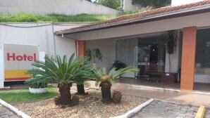 A hotel entrance with a sign, a paved walkway, and a small garden with palm trees. - Hotel Mime (Blumenau)