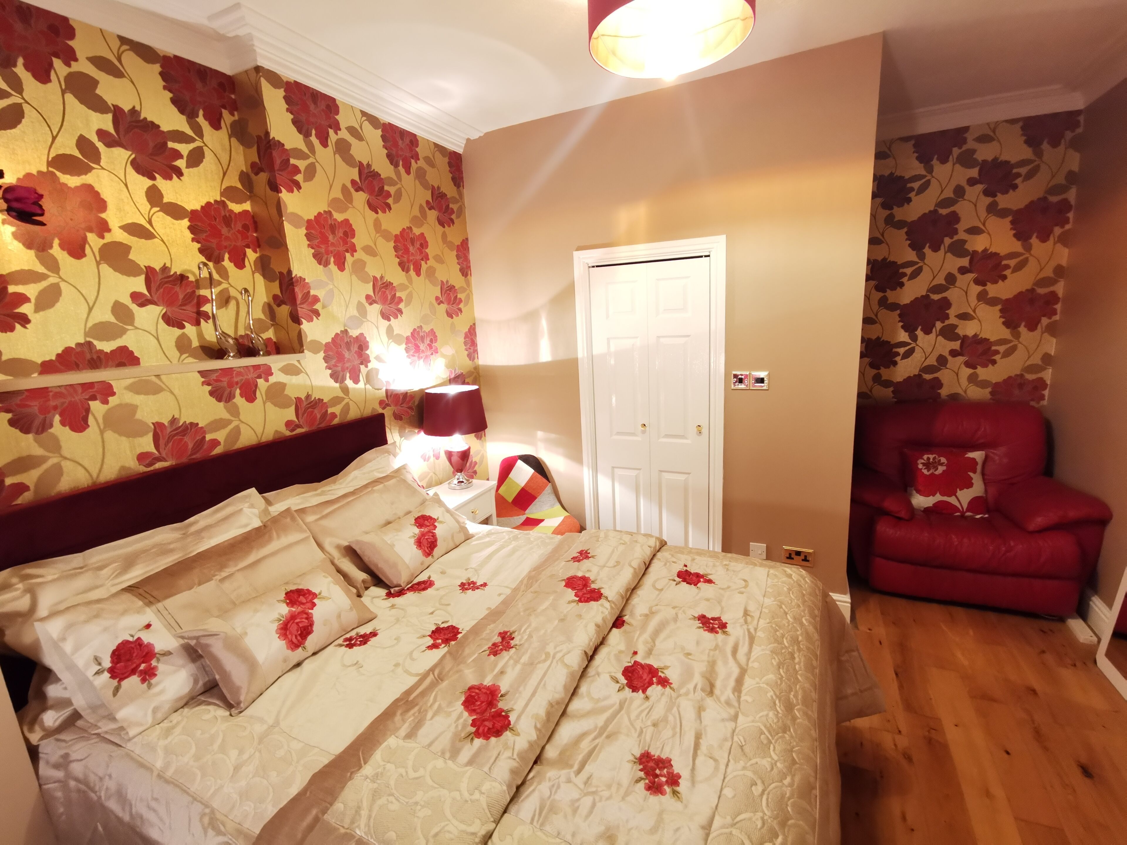 A bedroom with a bed, a red armchair, a nightstand with a lamp, and floral wallpaper.