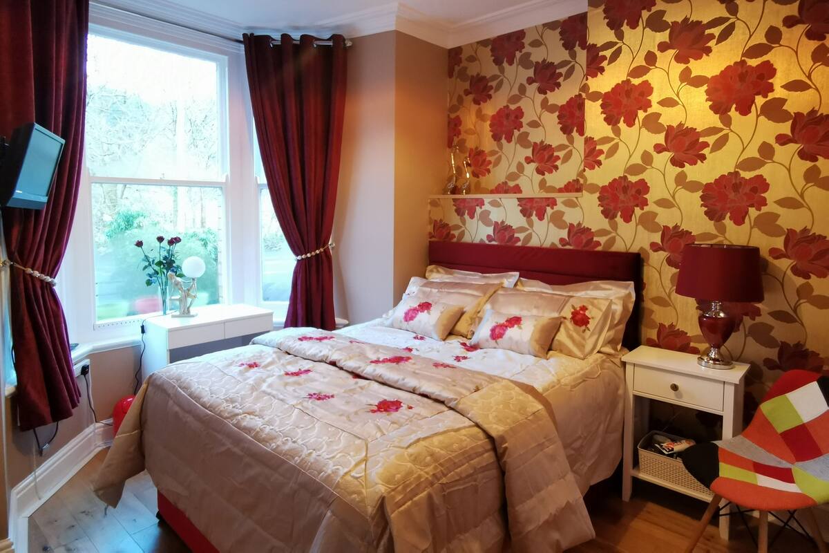 A bedroom with a bed, a nightstand, a lamp, a TV, and a window with curtains.
