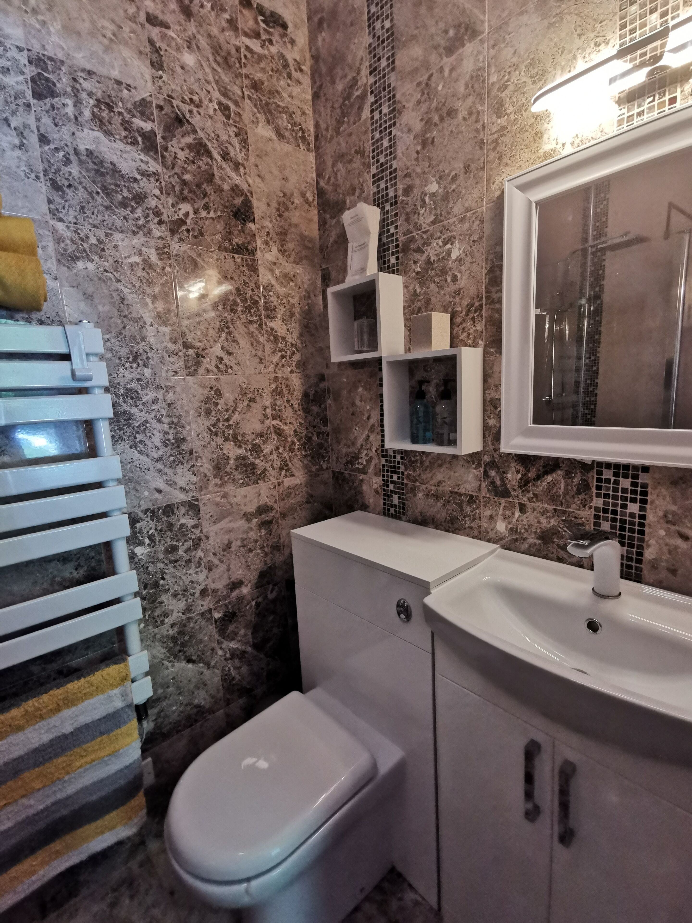 A bathroom with a marble wall, a white toilet, a sink with a mirror, and a towel rack.
