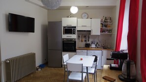 Une cuisine compacte équipée d’un réfrigérateur, d’un four à micro-ondes, d’un four et d’un lave-vaisselle. On y trouve également une table à manger avec des chaises, une horloge et une bouilloire.