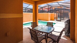 A sunlit patio with a glass table and chairs, a pool, and a view of a building with a red roof. - Paradise Palms-ru1074 (Kissimmee)