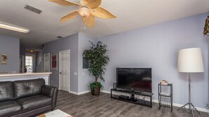 A living room with a dark leather couch, a red bowl on a white coffee table, a flat-screen TV on a black stand, a floor lamp, and a ceiling fan. - Windsor Palms-ru1025 (Kissimmee)