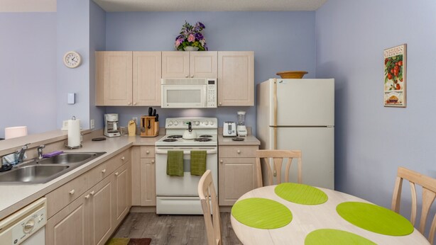 A compact kitchen with light wooden cabinets, a microwave, stove, and a refrigerator. A small dining table with a green placemat is visible. - Windsor Palms-ru1025 (Kissimmee)