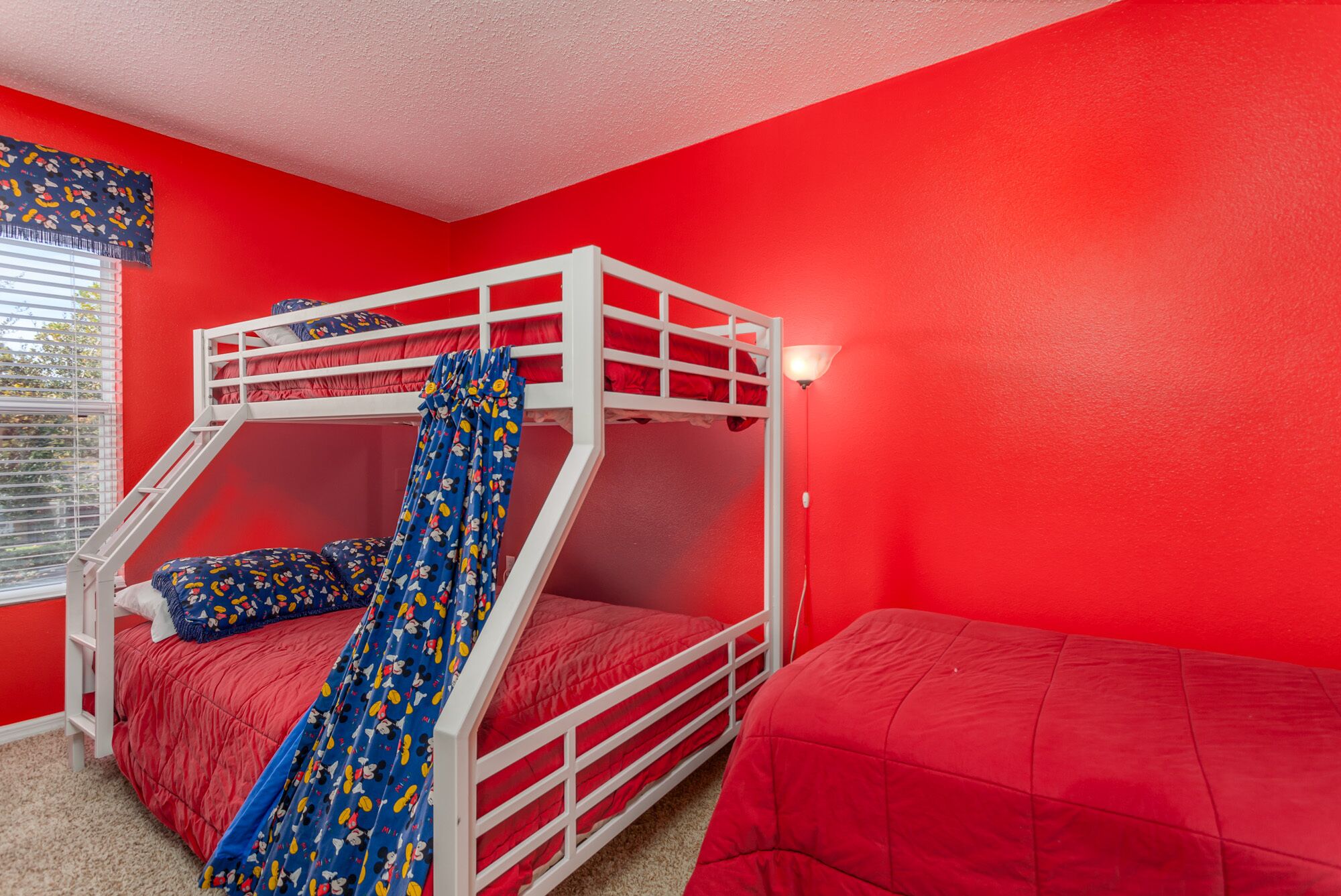 A bunk bed room with a red wall, a window with blinds, and a white ladder.