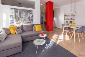 A modern living room with a grey sofa, yellow cushions, a small round table with a plant, and a dining area with a wooden table and chairs. - LOVELY APARTMENT SERRANOS TOWERS (València)