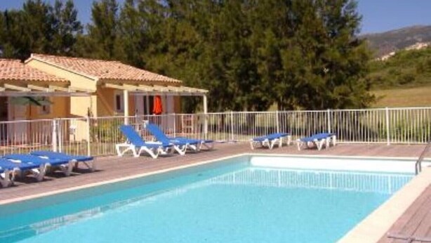 A swimming pool with blue lounge chairs, a covered area with a tiled roof, and a fence.