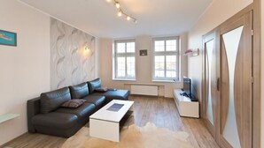 A modern living room with a leather sofa, a white coffee table, and a fur rug. There are two windows providing natural light, a radiator, and a television on a stand. - Gdańskie Apartamenty Targ Rybny Mołtawa (Gdansk)