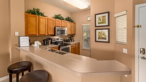 Une cuisine avec des meubles de rangement en bois, un four à micro-ondes en acier inoxydable, une cuisinière et un évier. On y trouve deux tabourets de bar et un tableau encadré accroché au mur.