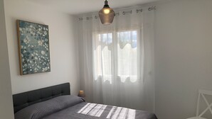 Une chambre à coucher avec un lit, une chaise, une lampe, un tableau et une fenêtre avec des rideaux.