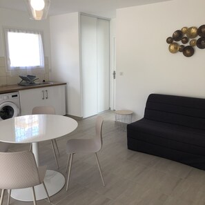 A modern living room with a black sofa, a round dining table with chairs, a kitchen area with a washing machine, and a sliding door leading to another room.