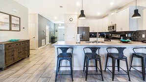 Une cuisine moderne avec des meubles blancs, un grand îlot central doté de tabourets, des appareils en acier inoxydable et un éclairage suspendu.