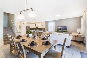 Un comedor con una mesa de madera preparada para seis personas, rodeada de sillas de madera. Se ve una cocina al fondo y un área de estar con un sofá y un televisor.
