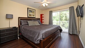 A bedroom with a wooden bed, a ceiling fan, a nightstand, a dresser, a TV mounted on the wall, and a window with curtains.