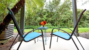 A patio with two blue chairs, a small table with a vase of flowers, and an air conditioning unit.